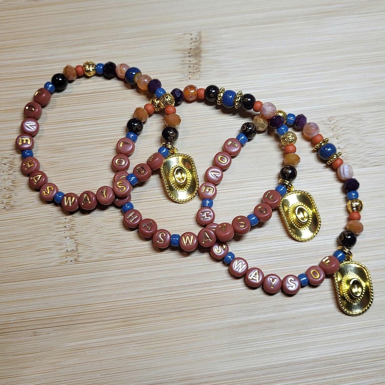 Peut inclure: Trois bracelets de perles avec des perles lettres mauves rondes &eacute;pelant ALWAYS, entrem&ecirc;l&eacute;es de petites perles bleues, noires, orange et dor&eacute;es. Chaque bracelet comporte une breloque chapeau de cowboy dor&eacute;e suspendue au cordon de perles.