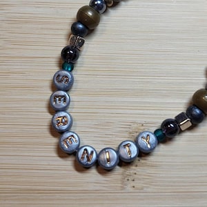 May include: A beaded bracelet with the word "SERENITY" spelled out in silver beads with gold lettering. The bracelet also features a mix of dark green, black, and gold beads. The bracelet is on a light brown wooden surface.