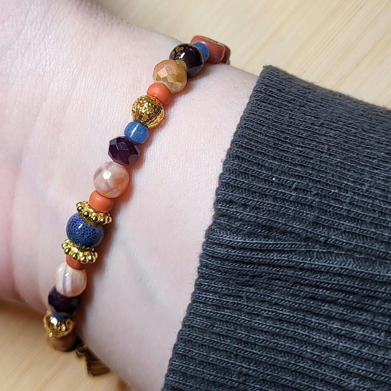 Peut inclure: Un bracelet de perles compos&eacute; de perles rondes et facett&eacute;es dans des tons orange, bleu, marron et cr&egrave;me. Le design int&egrave;gre des entretoises dor&eacute;es m&eacute;talliques et des perles textur&eacute;es, cr&eacute;ant un motif multicolore autour du poignet.