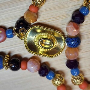 Peut inclure: Un collier avec un pendentif ovale dor&eacute; central dot&eacute; d'une bordure textur&eacute;e et d'un centre en relief. Le cordon est compos&eacute; de perles multicolores, notamment des perles orange facett&eacute;es, bleues lisses, violet fonc&eacute; et dor&eacute;es m&eacute;talliques.