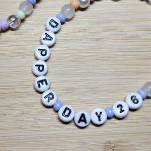 May include: A beaded bracelet featuring white circular beads with black lettering that spells out DAPPER DAY 26. The bracelet is constructed with a mix of translucent, light purple, and peach-coloured spherical beads interspersed between the lettered beads.