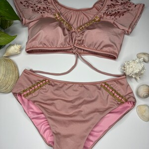 May include: A two-piece swimsuit in a dusty rose color. The top has short sleeves with a laser-cut design and gold and pink beaded trim. The bottoms are high-waisted with matching trim. Sunglasses are placed above the swimsuit.