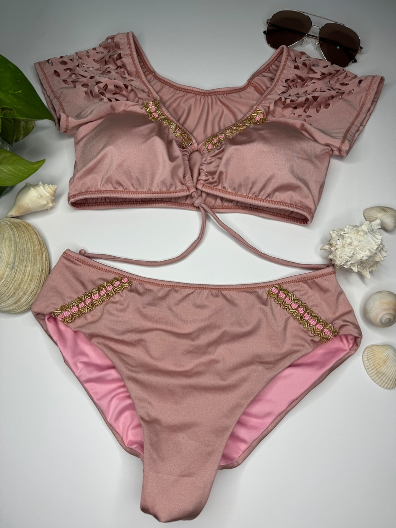 May include: A two-piece swimsuit in a dusty rose color. The top has short sleeves with a laser-cut design and gold and pink beaded trim. The bottoms are high-waisted with matching trim. Sunglasses are placed above the swimsuit.