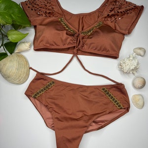 May include: A two-piece swimsuit in a warm brown hue. The top features laser-cut detailing on the sleeves and gold-colored embellishments. The bottoms have matching gold accents. Sunglasses and seashells are also visible.