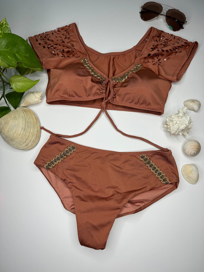 May include: A two-piece swimsuit in a warm brown hue. The top features laser-cut detailing on the sleeves and gold-colored embellishments. The bottoms have matching gold accents. Sunglasses and seashells are also visible.