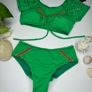 May include: A two-piece green swimsuit set. The top has laser-cut detailing on the sleeves and gold trim. The bottoms also feature gold trim. Sunglasses are placed above the swimsuit.