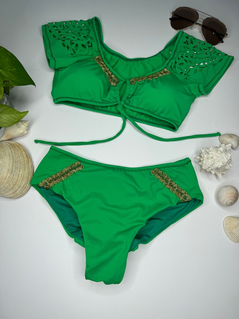 May include: A two-piece green swimsuit set. The top has laser-cut detailing on the sleeves and gold trim. The bottoms also feature gold trim. Sunglasses are placed above the swimsuit.