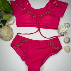 May include: A hot pink two-piece swimsuit set. The top has laser-cut detailing on the sleeves and gold trim. The bottoms also feature gold trim. Sunglasses are visible in the background.