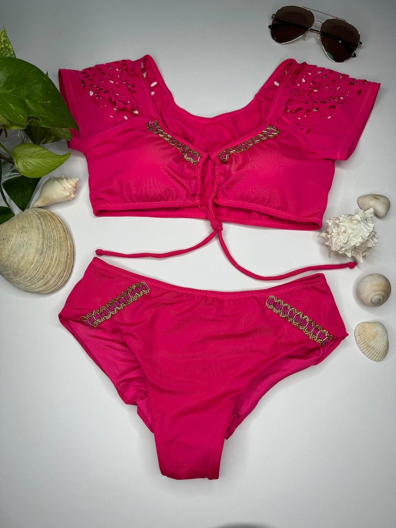 May include: A hot pink two-piece swimsuit set. The top has laser-cut detailing on the sleeves and gold trim. The bottoms also feature gold trim. Sunglasses are visible in the background.