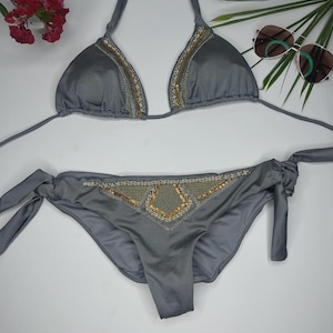 May include: A gray bikini set with gold sequin detailing. The triangle top and the bottoms have gold sequin accents. The bottoms have tie sides. Sunglasses and flowers are in the background.