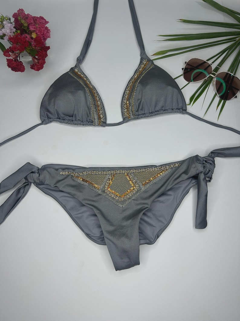May include: A gray bikini set with gold sequin detailing. The triangle top and the bottoms have gold sequin accents. The bottoms have tie sides. Sunglasses and flowers are in the background.