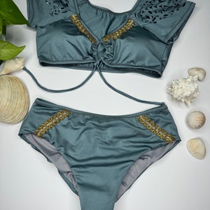 May include: A two-piece teal swimsuit set. The top has short sleeves with a leaf-like cutout design and gold-colored embellishments. The bottoms have matching gold embellishments. Sunglasses are visible in the background.