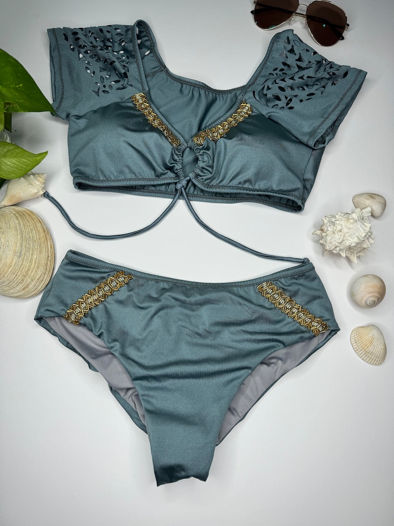May include: A two-piece teal swimsuit set. The top has short sleeves with a leaf-like cutout design and gold-colored embellishments. The bottoms have matching gold embellishments. Sunglasses are visible in the background.