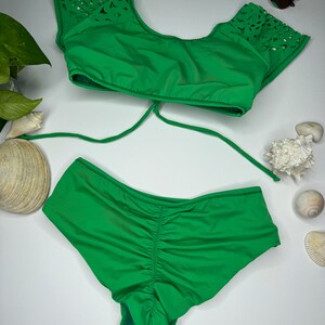 May include: A bright green bikini set with a top featuring laser-cut detailing on the sleeves and a ruched bottom. A pair of aviator sunglasses rests nearby. The swimwear is displayed on a white surface, suggesting a summer or beach theme.