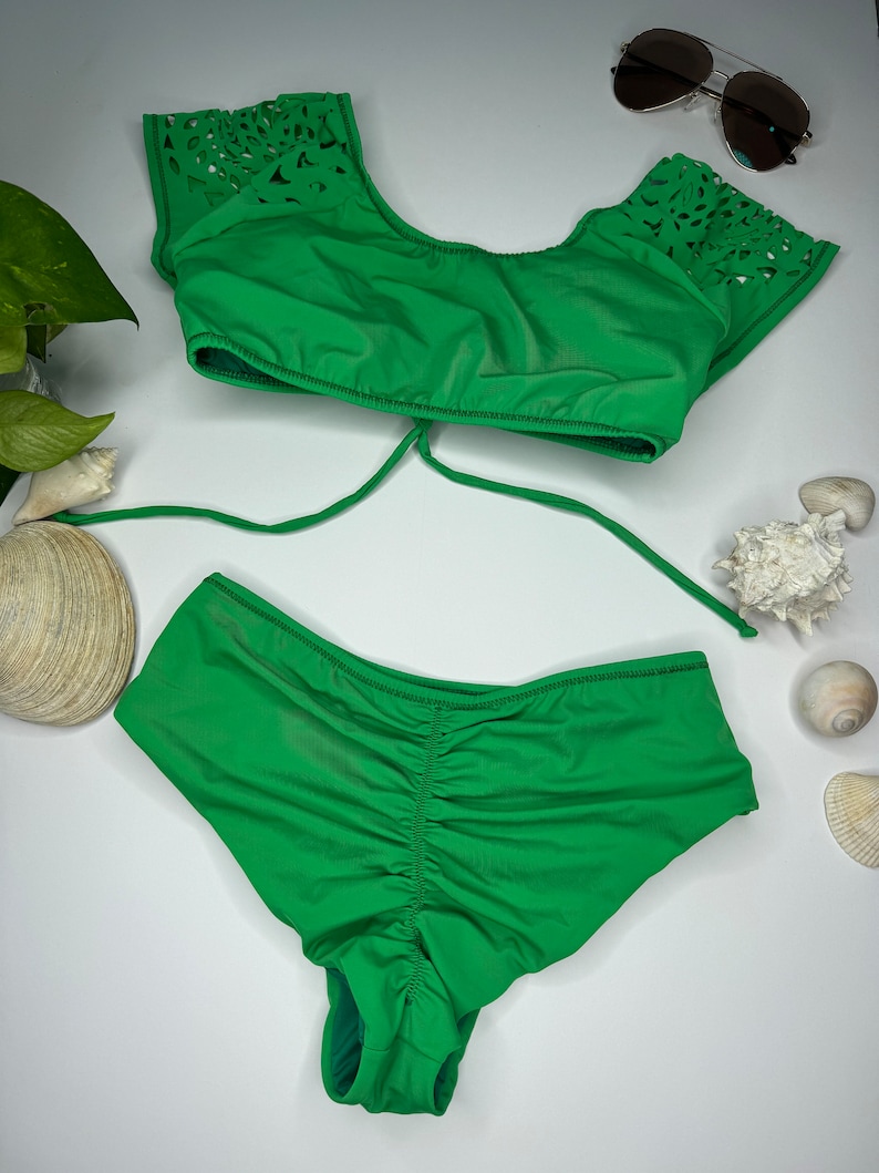May include: A bright green bikini set with a top featuring laser-cut detailing on the sleeves and a ruched bottom. A pair of aviator sunglasses rests nearby. The swimwear is displayed on a white surface, suggesting a summer or beach theme.