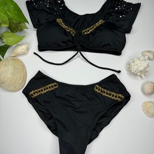 May include: Black two-piece swimsuit with gold chain accents. The top has a tie closure and cut-out detailing on the sleeves. The bottoms feature gold chain trim on the sides. Sunglasses are also visible.