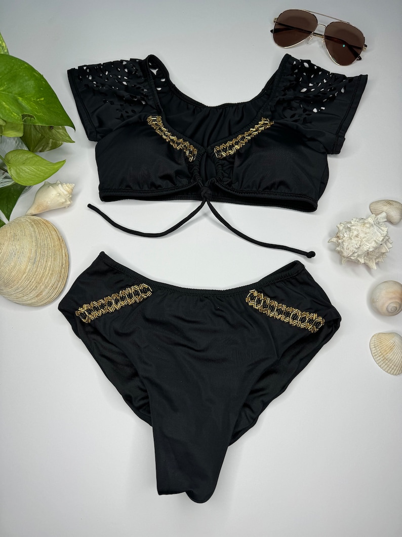 May include: Black two-piece swimsuit with gold chain accents. The top has a tie closure and cut-out detailing on the sleeves. The bottoms feature gold chain trim on the sides. Sunglasses are also visible.