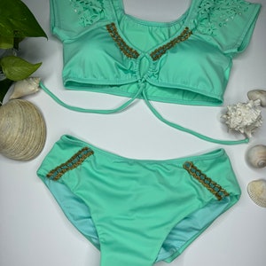 May include: A mint green bikini set with a matching top and bottom. The top features laser-cut detailing on the sleeves and gold and turquoise beading along the neckline. The bottoms have similar beading on the sides. Sunglasses are placed above the bikini.
