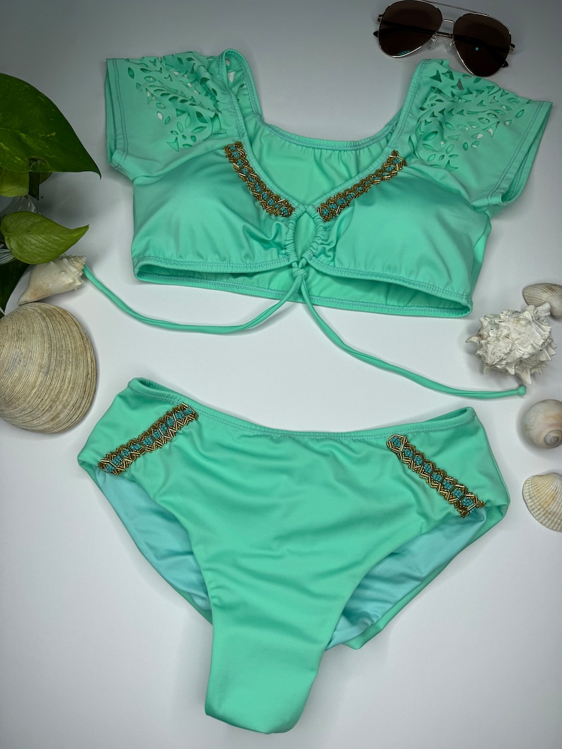 May include: A mint green bikini set with a matching top and bottom. The top features laser-cut detailing on the sleeves and gold and turquoise beading along the neckline. The bottoms have similar beading on the sides. Sunglasses are placed above the bikini.