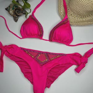 May include: A vibrant pink bikini set. The triangle top and low-rise bottoms feature decorative beading along the edges and center. The bikini top has halter straps, and the bottoms have side ties. A straw hat is in the background.