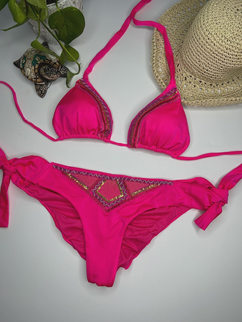 May include: A vibrant pink bikini set. The triangle top and low-rise bottoms feature decorative beading along the edges and center. The bikini top has halter straps, and the bottoms have side ties. A straw hat is in the background.