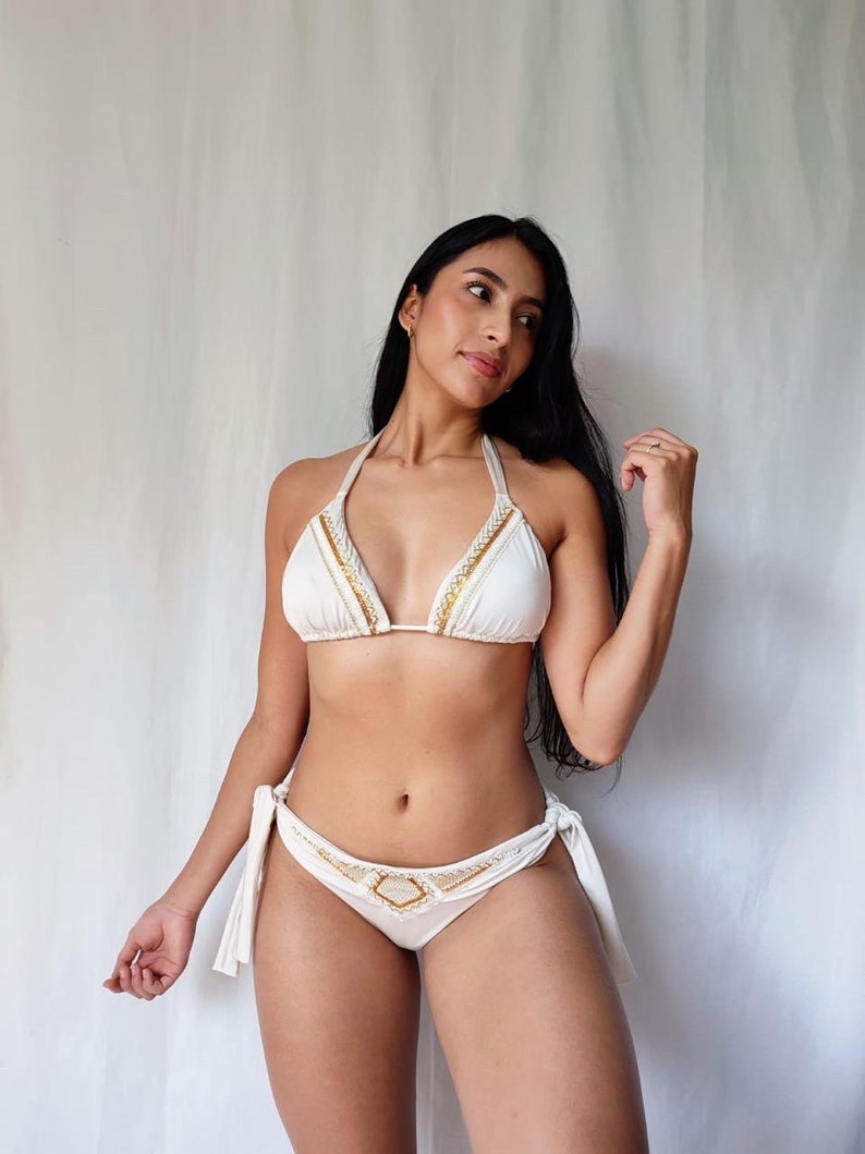 May include: White bikini with gold and cream-colored trim. The triangle top and low-rise bottoms feature decorative accents. The bottoms have tie sides. The bikini is made of a soft, lightweight material.
