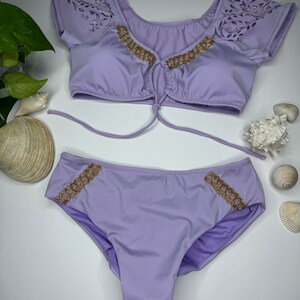 May include: A two-piece lavender swimsuit set. The top has short sleeves with a laser-cut design and gold trim. The bottoms also feature gold trim. Sunglasses are visible in the background.