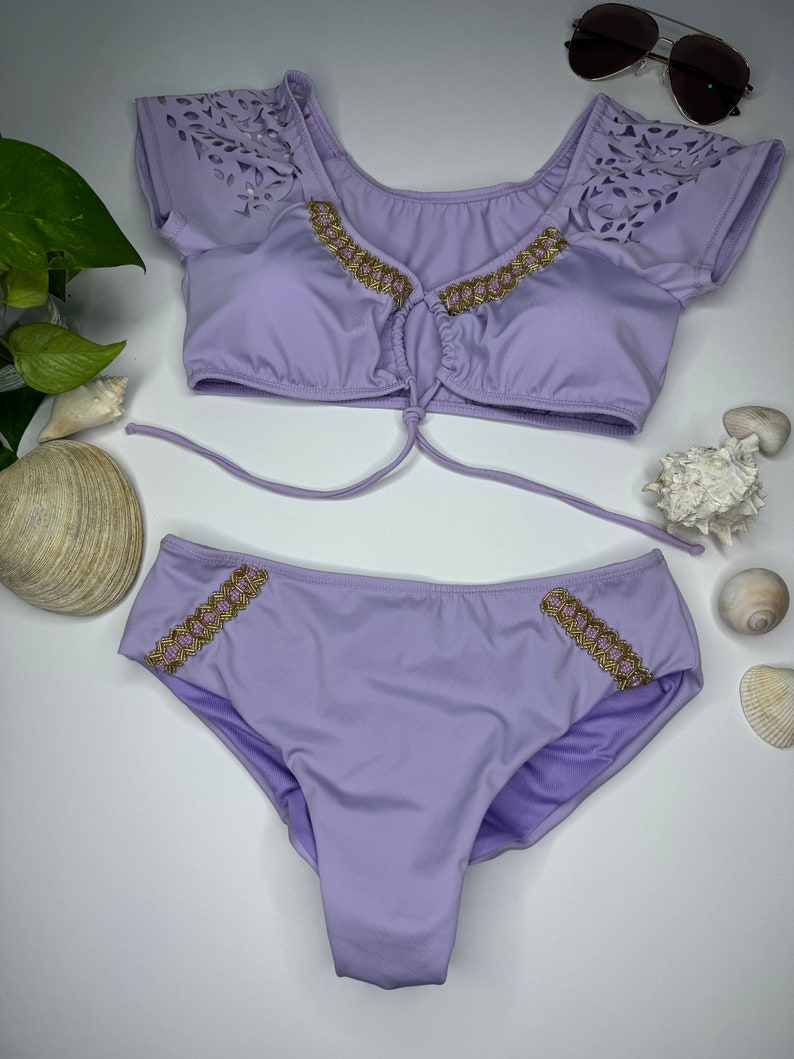 May include: A two-piece lavender swimsuit set. The top has short sleeves with a laser-cut design and gold trim. The bottoms also feature gold trim. Sunglasses are visible in the background.