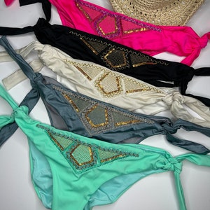 May include: A collection of five bikini bottoms in vibrant colors: hot pink, black, cream, gray, and mint green. Each features a decorative sequined design and tie sides. A straw hat is visible in the background.