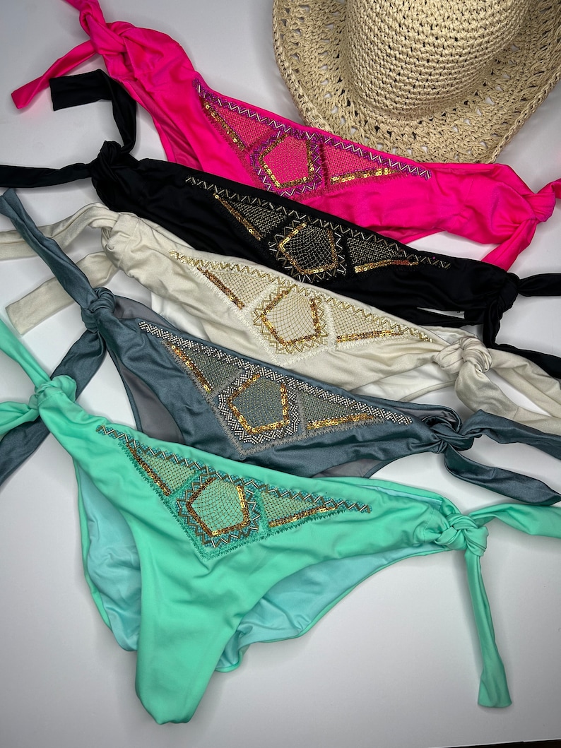 May include: A collection of five bikini bottoms in vibrant colors: hot pink, black, cream, gray, and mint green. Each features a decorative sequined design and tie sides. A straw hat is visible in the background.
