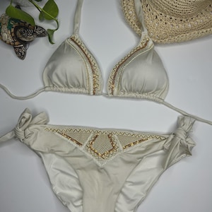 May include: Cream-colored bikini set with intricate beaded and embroidered detailing. The triangle top and low-rise bottoms feature gold and white accents. A straw hat is visible in the background, suggesting a beach or vacation theme.