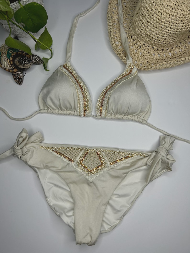 May include: Cream-colored bikini set with intricate beaded and embroidered detailing. The triangle top and low-rise bottoms feature gold and white accents. A straw hat is visible in the background, suggesting a beach or vacation theme.