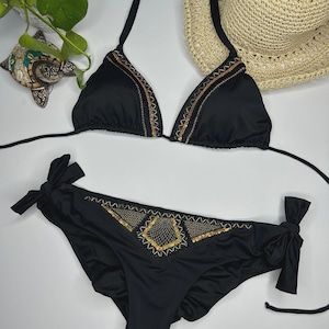 May include: Black bikini set with triangle top and low-rise bottoms. The bikini top and bottoms are adorned with gold and bronze embellishments. A straw sun hat is visible in the background.