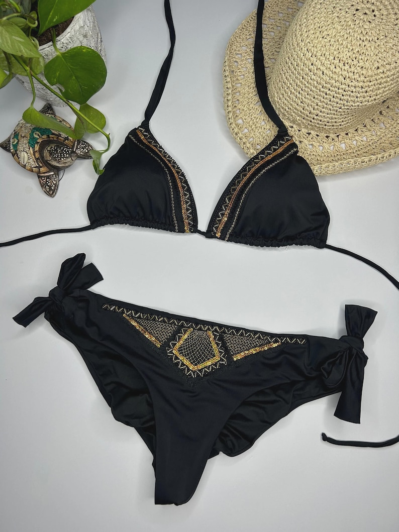 May include: Black bikini set with triangle top and low-rise bottoms. The bikini top and bottoms are adorned with gold and bronze embellishments. A straw sun hat is visible in the background.