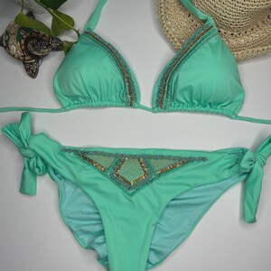 May include: A mint green bikini set with triangle top and tie-side bottoms. The edges of the top and the center of the bottoms are embellished with gold and teal beading. A straw hat is in the background.