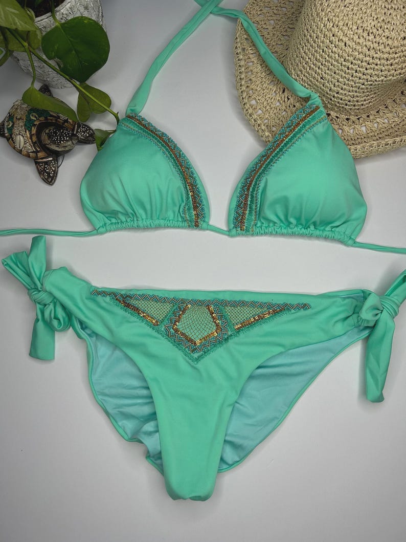 May include: A mint green bikini set with triangle top and tie-side bottoms. The edges of the top and the center of the bottoms are embellished with gold and teal beading. A straw hat is in the background.