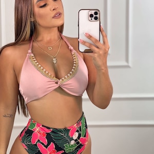May include: A two-piece swimsuit featuring a light pink halter top with gold embellishments and a high-waisted bottom. The bottom has a black background with a vibrant floral pattern in pink and green.