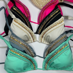 May include: A collection of triangle bikini tops in various colors, including pink, black, white, teal, and seafoam green. Each top features decorative gold and silver beading along the edges. A straw sun hat is partially visible in the background.