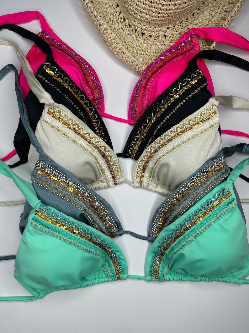 May include: A collection of triangle bikini tops in various colors, including pink, black, white, teal, and seafoam green. Each top features decorative gold and silver beading along the edges. A straw sun hat is partially visible in the background.
