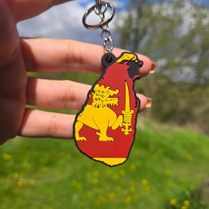 May include: A keychain shaped like the island of Sri Lanka with the Sri Lankan flag on it. The flag features a lion holding a sword on a red background with a yellow border.