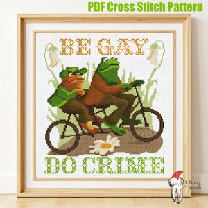 May include: Cross stitch pattern featuring two frogs riding a bicycle with the text "Be Gay Do Crime" in green and yellow letters.