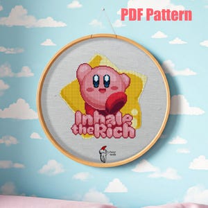 May include: A cross-stitch pattern featuring a pink Kirby character from the video game series Kirby, holding a star with the text "Inhale the Rich" on a white background.