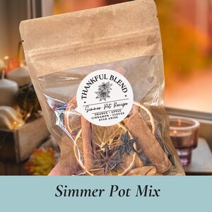 May include: A clear bag filled with a simmer pot recipe, including orange slices, cinnamon sticks, and star anise. The bag has a label that reads "Thankful Blend" and "Simmer Pot Recipe". The image is set against a warm, autumnal background.