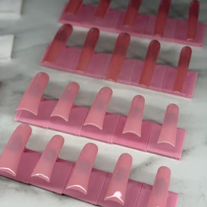 May include: Rows of pink acrylic nail tips arranged on pink holders. The tips are a gradient of pink, transitioning from a darker shade at the top to a lighter shade at the base. The nails are long and square-shaped, ready for application.