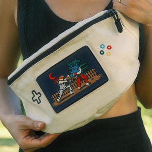May include: A cream-colored canvas fanny pack featuring a rectangular patch with a pixelated fighting game scene. The patch shows two characters in combat, a red crescent moon, and a fence. A small white cross and four tiny colored flower icons are embroidered.