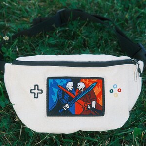 May include: A cream-coloured bum bag featuring a central rectangular patch depicting two figures with swords in blue and red settings. The bag includes an embroidered black cross-shaped icon on the left and four small coloured circles on the right.