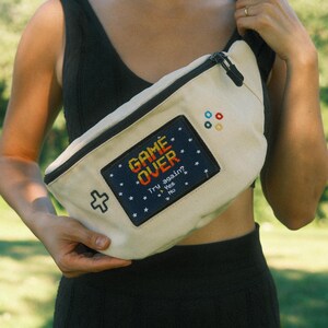 May include: A cream-colored canvas fanny pack featuring a black rectangular patch with the pixelated text GAME OVER, Try again? Yes No. The bag includes a black zipper, a white cross-shaped icon, and four small colored circles on the front.