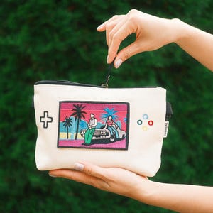 May include: A cream-colored zippered pouch featuring an embroidered rectangular patch of a car with two figures under palm trees against a sunset background. The pouch includes a black zipper, a cross-shaped icon, and four small colored circles.