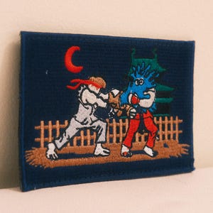 May include: A rectangular embroidered patch on a dark blue background featuring a white-clad martial artist with a red headband fighting a blue creature. The scene includes a brown wooden fence, a green pagoda-style building, and a red crescent moon.
