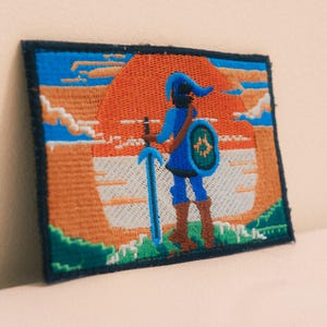 May include: A rectangular embroidered patch featuring a figure in blue clothing standing with a sword and shield against a large orange sun. The scene includes a blue sky, white clouds, and green terrain, all bordered by a dark navy blue stitched edge.